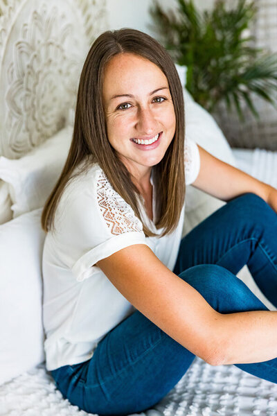 Woman in white shirt and brown hair sits on a bed with legs up and arms hugging them.
