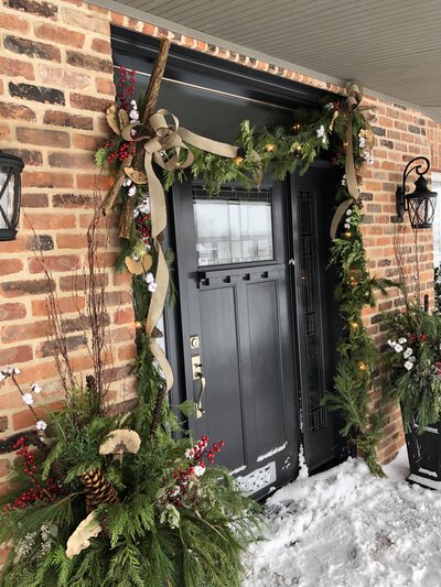 Custom outdoor exterior decorations around a black door with cedar, pine tree branches and ribbon by Helena's gardening