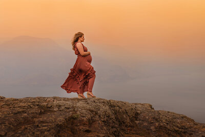 A pregnant women wearing a brown dress stands on the edge of a cliff cradling her belly with a sunset smokey sky in the background. Captured by Brittany Danielle