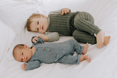 Toddler gently holding his newborn baby brother, both lying on a soft cream blanket. A quiet, loving moment captured by Sophie Louise for Nostalgia.