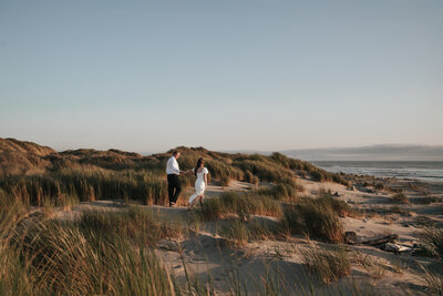 Bride and groom walking through scenic sand dunes in Florence, Oregon, surrounded by tall coastal grass and golden hour ocean views during a romantic Oregon Coast engagement session.