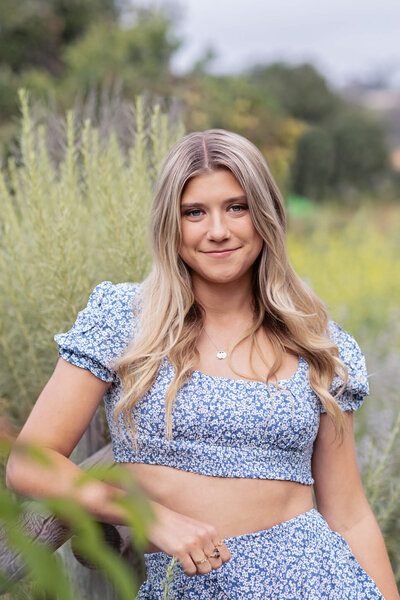 A blonde young woman wearing a blue UCLA sweatshirt giving the "fours up" sign while standing in a field of yellow flowers