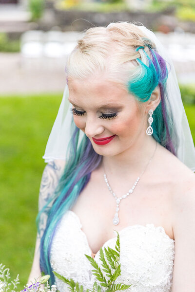 Bride with purple and blue hair looking down at her flowers with a red lip.