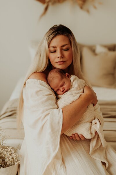 Mother softly rests her head on her newborn whom she is holding close to her chest. They are sitting in a elegant photo studio