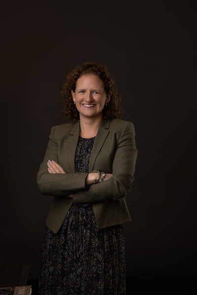 corporate headshot of business woman with long dress and dark olive green jacket on a dark background
