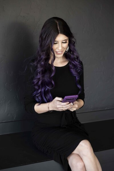 Laura is wearing a black dress, sitting on a black bench against a gray wall. She is holding a smartphone with both hands, smiling down at it. The phone has a purple case, which matches her long purple hair, which flows down past her shoulders to her elbows.