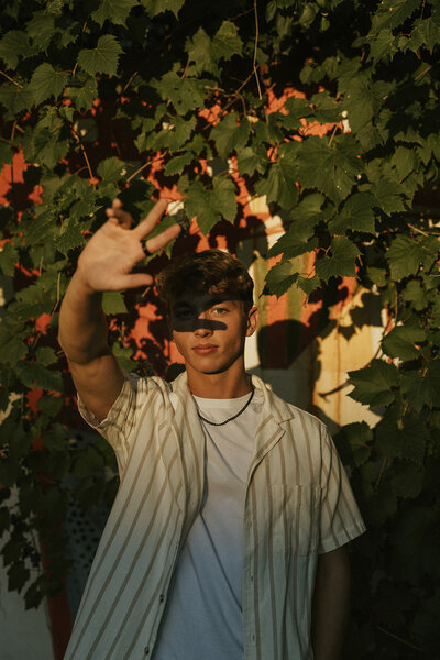 A senior boy standing in front of a wall of greenery as the last light hits his face and casts a shadow over it. 