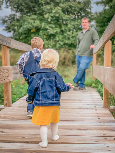 Wooden bridge at the end of it dad. He is recognizable but blurry. In the front of the picture. Two kids running towards him. Both dressed in denim.  Motion of them running very visible. Family Photography.