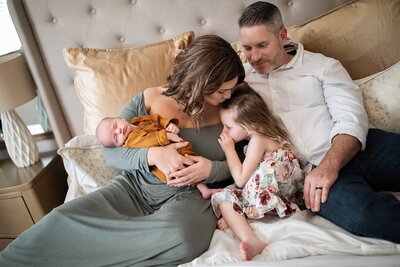 Family sitting on bed with mom holding baby and kissing big sisters head for Orlando Newborn Photos