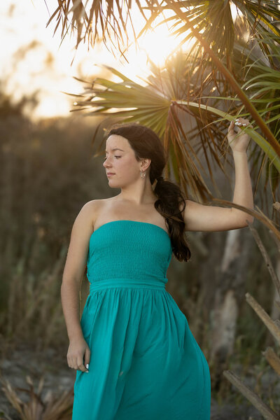 A senior girl in a flowing turquoise dress poses gracefully under palm trees at sunset. The golden light illuminates her face as she gently holds onto a palm frond, creating a soft, dreamy aesthetic perfect for a coastal-inspired senior photoshoot.