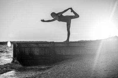 Black and white photo of woman standing on ocean dock in a yoga pose wearing a black yoga onesie.