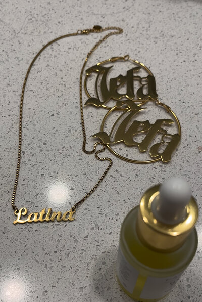 Gold Latina name plate necklace, Jefa gold hoops, and gold bottle.