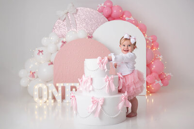 Toddler holding on to a giant cake prop with bows and arches in the background in pink and white.