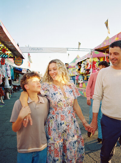 Mother walks with husband and middle son down a parade of fair games at sunset.