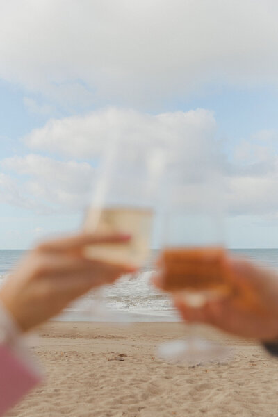 Two people toasting with wine glasses on a beach in San Clemente, California. The ocean waves and a cloudy sky are in the background, creating a serene and celebratory atmosphere. Shindig Social promotes authentic gatherings and meaningful connections.