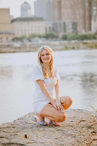 Blonde Girl Senior Kneeling in white dress on Rock overlooking water. 