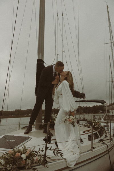 A couple shares a kiss on a sailboat, surrounded by flowers, against a moody sky and calm waters.