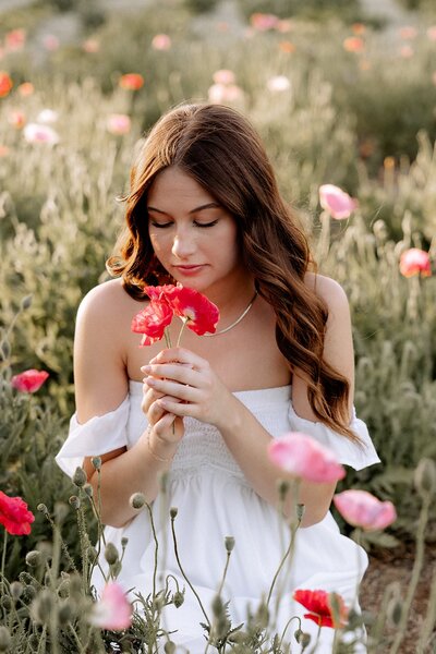 mom in a elegant navy blue dress sitting in a grass field with a pathway lined with trees behind her. She is smiling down at her older daughter as she hugs both girls .