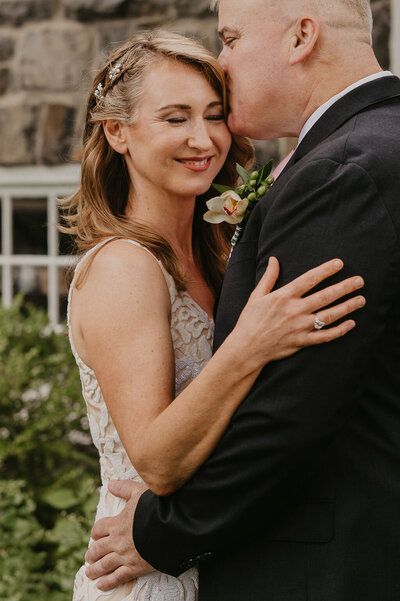 A bride in a lace dress embraced by her husband closes her eyes as she is kissed on the head by her groom