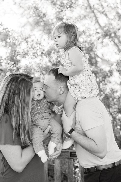black and white photo of a fun family with the little girl on dad's shoulders while mom and dad are kissing their baby boy's cheeks