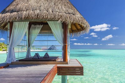 Thatched-roof cabana with white curtains overlooking clear turquoise water on a tropical island.