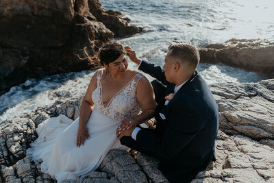 Bride and groom are sitting on a rock above the ocean groom is  tucking bride's hair behind her ear and they are holding hands
