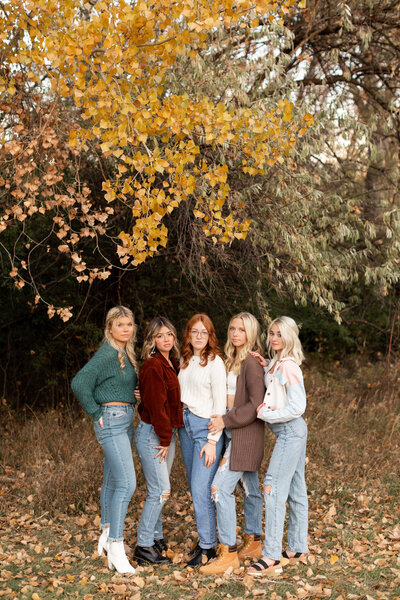 Five high school seniors posing together with hands on each other underneath a tree changing color and leaves on the ground.