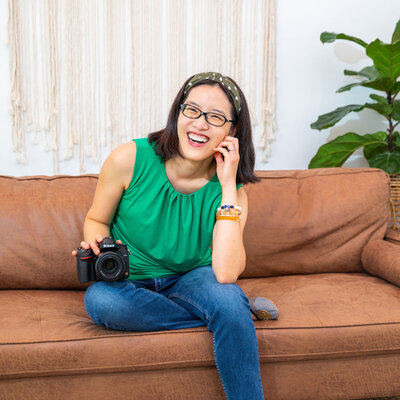 An Asian woman sitting on a brown couch with her DSLR and smiling.