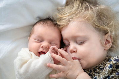 Toddler sucks thumb while lying beside a newborn baby on a bed during a cozy newborn photography session in Munich.