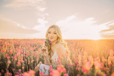 woman with light hair sitting in a field of pink flowers at sunset
