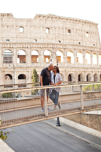 A couple standing on a bridge with the Colosseum in the background. Taken by Rome Photographer, Tricia Anne Photography.