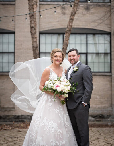 groom and bride with billowing veil in front of brick building
