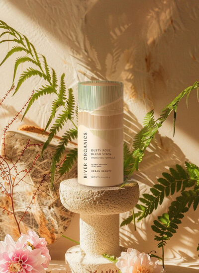 River Organics' Dusty Rose Blush Stick is displayed on a stone pedestal surrounded by lush green ferns. The eco-friendly packaging, featuring soft green and beige tones, reflects the brand’s commitment to vegan beauty and natural ingredients. Sunlight filters through the leaves, creating a warm, earthy ambiance that highlights the product’s organic and hydrating formula.