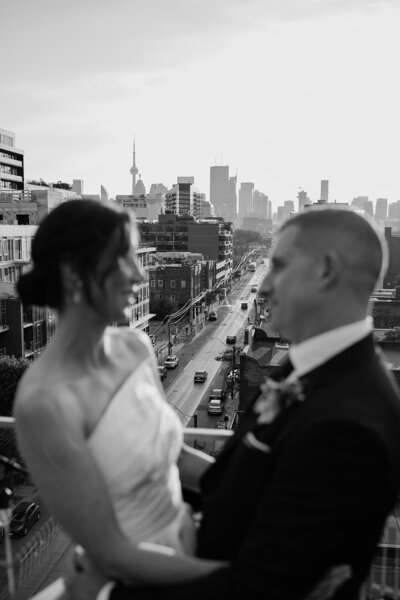 A couple in formal attire stands on a rooftop at the Broadview Hotel, gazing at each other amidst a cityscape with a prominent tower in the background. Captured in timeless black and white by Toronto wedding photographer, Jennifer van Son.
