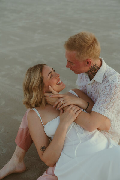 Engaged couple sitting on the beach at sunset, gazing into each other's eyes with joyful smiles, wearing white and pink outfits during a Dayton Beach proposal session.