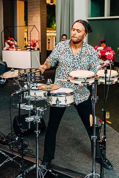 Male percussionist playing a multi-piece drum kit with focused expression during an elegant indoor event.