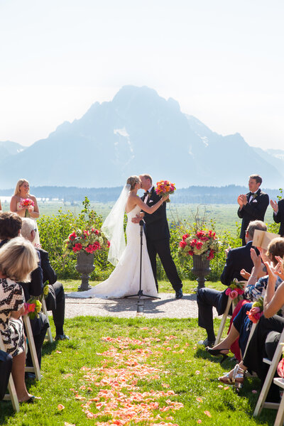 Jackson Hole Wedding Ceremony with Grand Teton Backdrop. A bride and groom stand at the altar, surrounded by guests during their outdoor ceremony with vibrant pink and orange floral arrangements framing the scene.