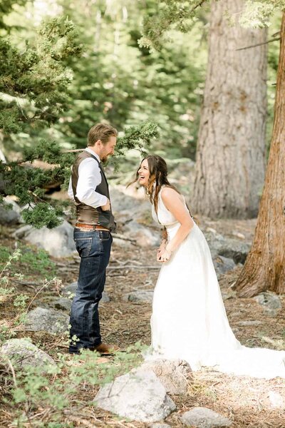 A bride laughs while she shares a first look with her groom while in forest surrounded by trees. 