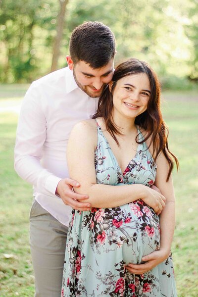 Pregnant woman smiling off camera, with both hands on her stomach. Her partner stands behind her, looking over her shoulder at her belly.