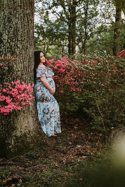 Pregnant mom in blue floral dress standing next to a tree in a flower garden holding her pregnant belly in Brighton Maryland.