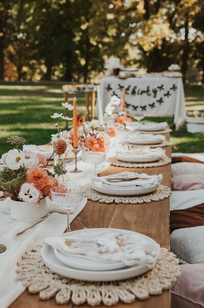 Outdoor celebration with wooden picnic tables decorated with orange and white florals, candles, and elegant tableware, styled by Yours Truly, a wedding planner and designer in New Jersey.
