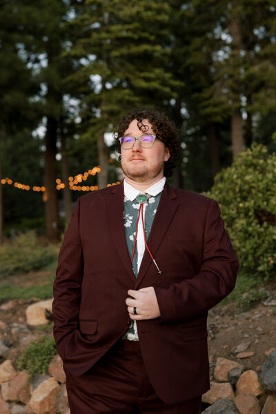 Lake Tahoe Wedding Photographer Greg wearing a maroon suit on his wedding day, he looks off at the sunset as he holds his suit jacket with his other hand in his pocket. There are some pine trees behind him