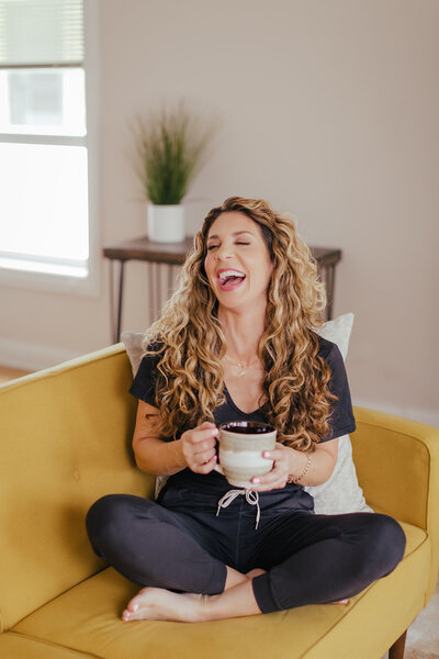 Curly haired woman with light skin sitting on a yellow couch in sweats and a t-shirt. She is holding a large mug of coffee and laughing hard.