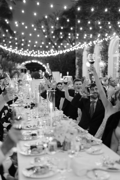 Guests raising their glasses in celebration at a long banquet table lit with romantic string lights.