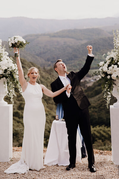 Bride and groom celebrating. Bride raises bouquet in the air. Carmel valley in the background
