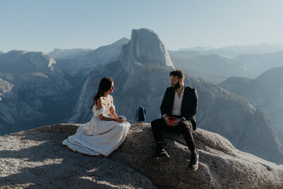 bride booping groom's nose with frosting on her finger from wedding bundt cake that is on their picnic blanket in front of them there are beige grasses and shrubs in the background