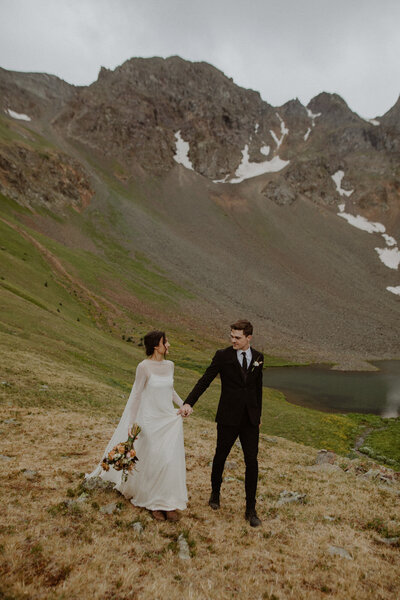 Eloping couple walk in front of an alpine lake while holding hands