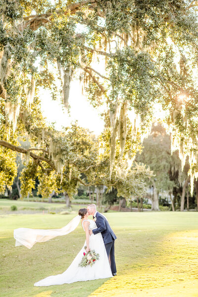 Bride and groom kiss while viel flies. They are standing under Spanish moss with the sin shining through in Charleston, SC
