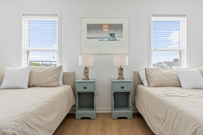 Beach-inspired guest room with crisp white walls, two small beds, light blue vintage nightstands, eclectic lamps, and views of the outdoors.
