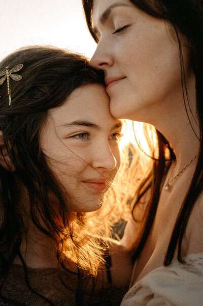 Older sister leans down to wrap her arm around her little sister's shoulder as they look off into the sunset. 